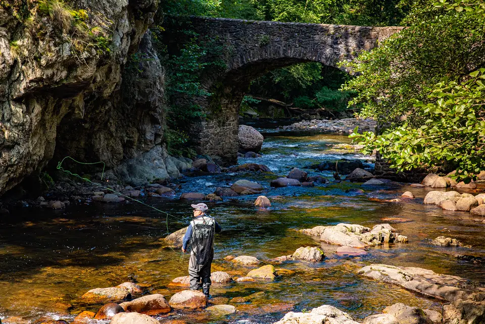 Pescador con vadeador en un río de aguas claras junto a un puente de piedra