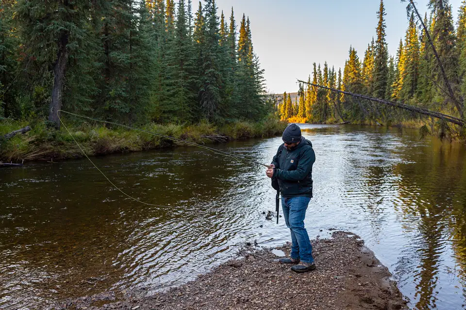 Pescador revisa su equipo en la orilla de grava de un río rodeado de bosque