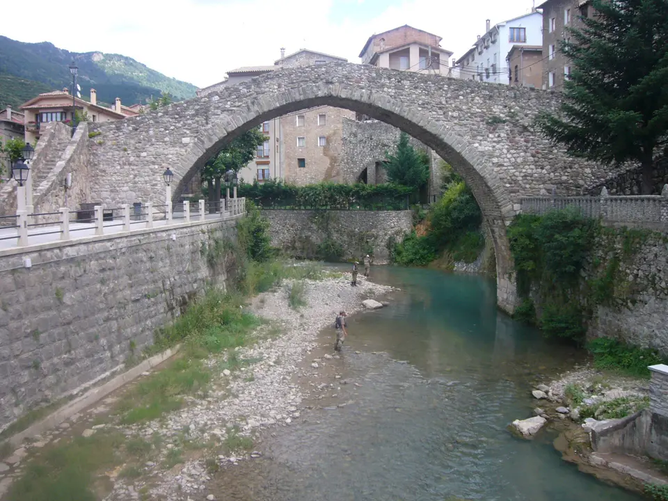 Puente medieval de piedra sobre un río de aguas claras con personas en el cauce