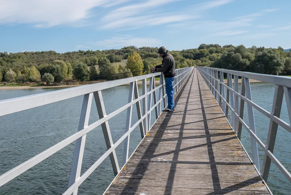 Pescador en una pasarela metálica sobre un embalse rodeado de arbolado